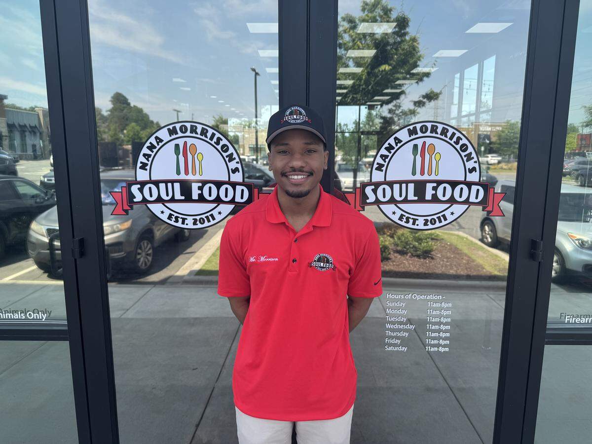 A medium shot of a young man smiling and standing in front of the double glass doors of a restaurant. He is wearing a red polo shirt with an embroidered logo on the chest and a black baseball cap. The glass doors behind him feature two circular logos that read “NANA MORRISON’S SOUL FOOD EST. 2011” flanking him, with the restaurant’s hours of operation printed in white text on the lower right pane.