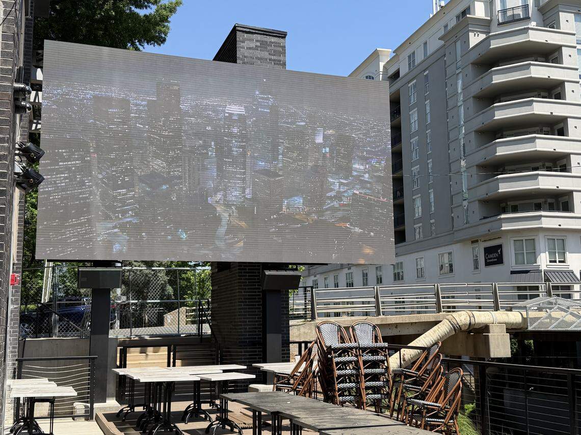An outdoor patio area with stacked bistro chairs and black tables, dominated by a massive outdoor LED screen displaying a nighttime city skyline.