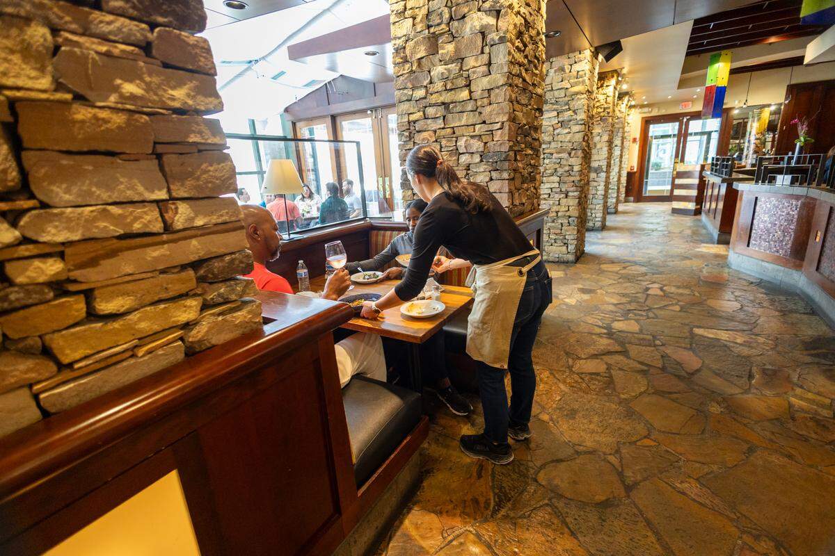 An interior shot of the restaurant’s dining area featuring rustic stone pillars and slate tile flooring. A server is leaning over a wooden booth table to serve a group of diners who are enjoying drinks and appetizers.