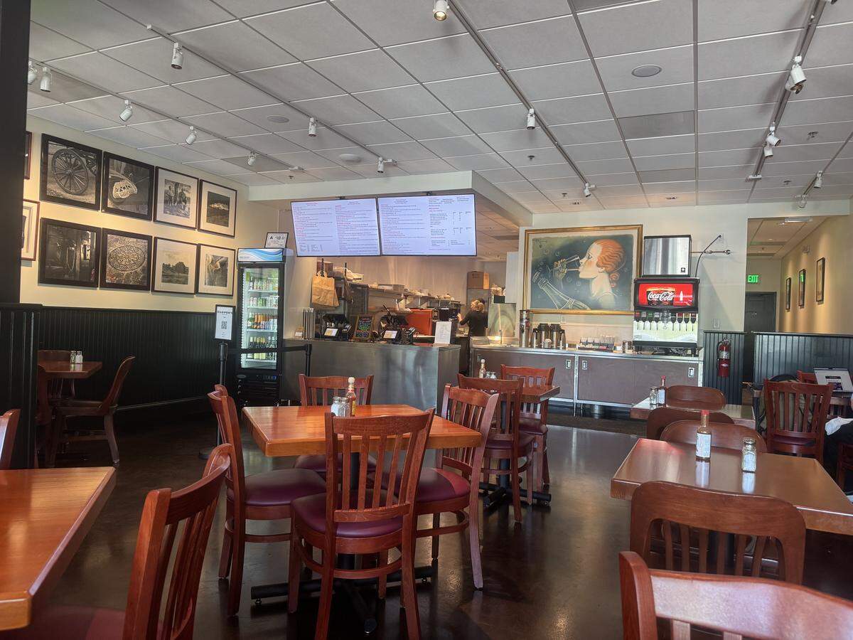 A medium-angle interior shot of a casual restaurant dining room. The room has multiple wooden tables with matching wooden chairs with red seats. In the background, there is a counter area with a soda machine, a drink cooler, and two large digital menu boards hanging above the counter. The walls are decorated with framed black and white pictures and a large framed painting of a woman with red hair and a red dress playing a trumpet.