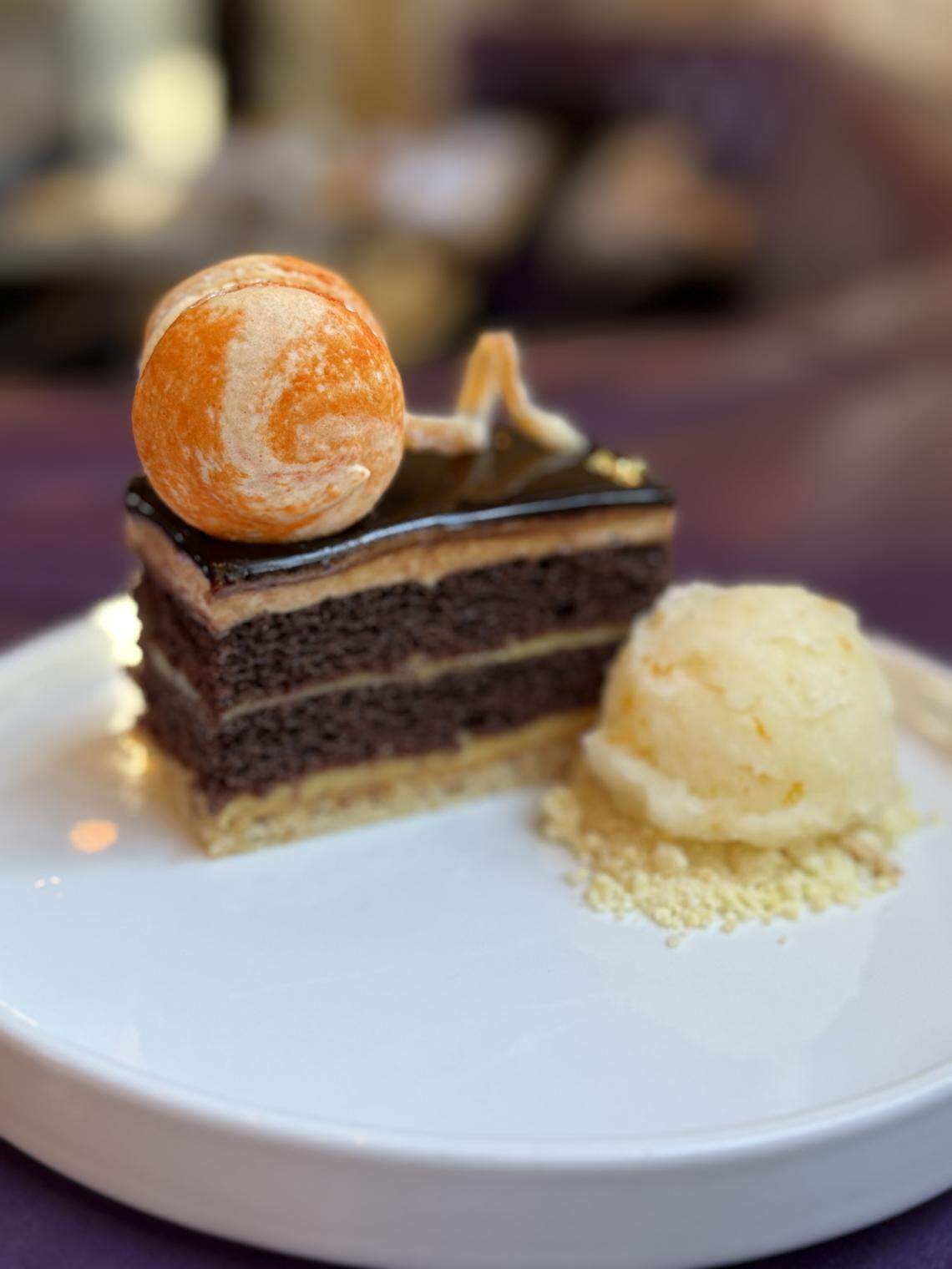 A square, layered chocolate cake with a dark glaze, topped with a sphere of orange and white meringue or chocolate, served on a white plate with a scoop of light yellow sorbet and crumbled topping. The dessert is likely the Petits Gateaux from Supperland.