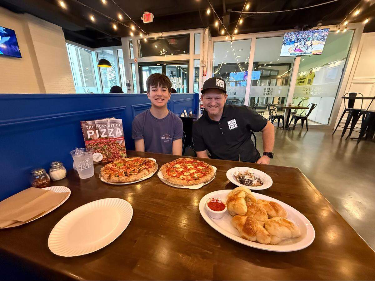 A young person and a chef sit together at a long wooden table, posing for a photo. The table is filled with a feast: two large pizzas, a massive plate of garlic knots with marinara sauce, a plate of cannolis, and a “Pursuit of Pizza” cookbook.
