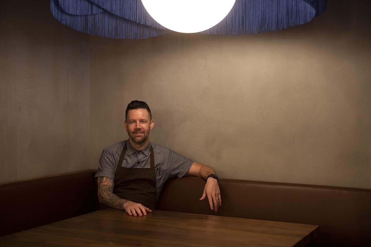 Interior shot of a chef sitting in a cozy, upscale restaurant booth under a soft, circular light fixture. The chef is smiling and appears relaxed at a dark wooden table.