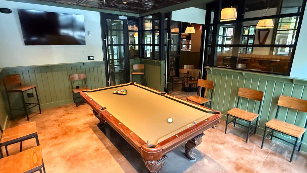 A dedicated billiards room within the pub. A tan-felted pool table sits in the center of the room, which features sage green wainscoting and a dark tin-style ceiling. Wooden chairs and barstools are placed along the walls for spectators. Large glass-paned partitions separate this area from the main dining room while maintaining an open feel.