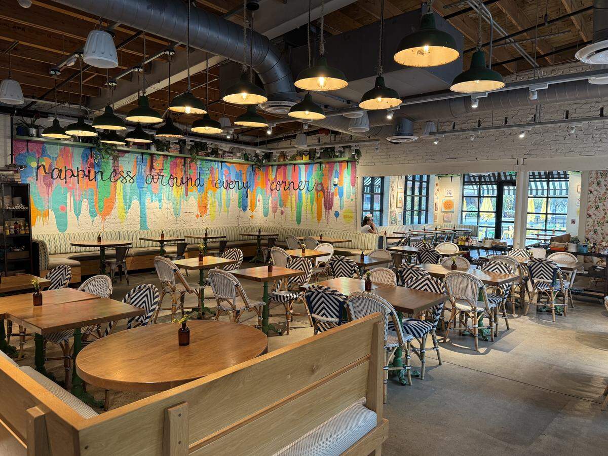 A wide interior view of a bright and cheerful restaurant dining room with an eclectic, bohemian style. The back wall features a colorful mural with the text “HAPPINESS AROUND EVERY CORNER.” The room is filled with wooden tables and chairs with navy blue and white patterned wicker backs and seats.