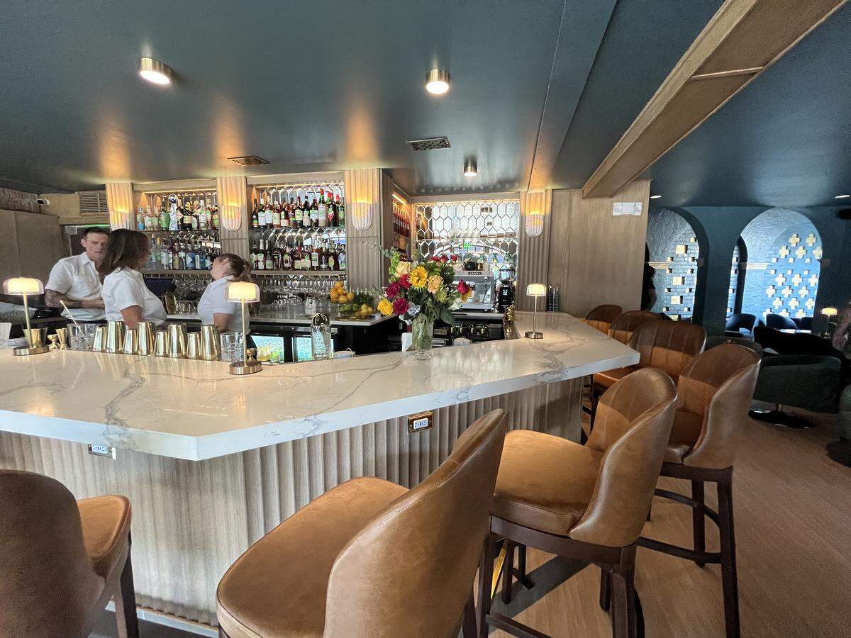 The elegant interior of an upscale bar with an Art Deco feel. A long, white marble bar is the centerpiece, with a row of brown leather bar stools in front. The back bar features three illuminated, arched alcoves filled with bottles. Bartenders are seen preparing drinks, and a large bouquet of flowers sits on the counter.