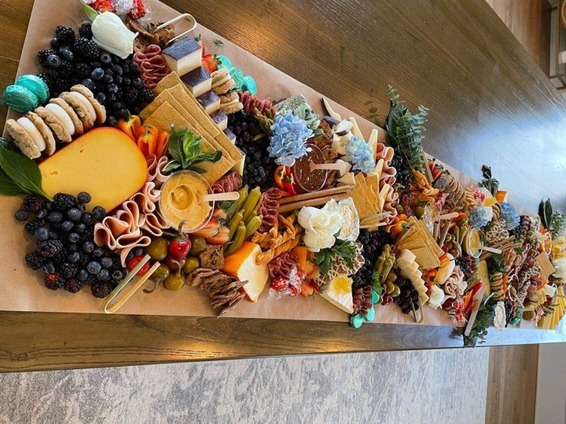 A massive charcuterie board, overflowing with a variety of cheeses, crackers, fruits, and meats, is displayed on a long wooden table. The arrangement features several large wedges of cheese, bowls of dips, and edible flowers, suggesting a festive and abundant gathering.