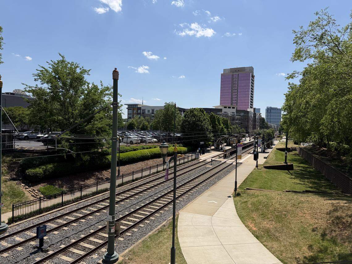 A high-angle view of a light rail station platform and tracks stretching toward the Charlotte skyline. Lush green trees and a parking lot line the tracks, with a distinctive tall building featuring purple reflective glass visible in the background under a clear blue sky.