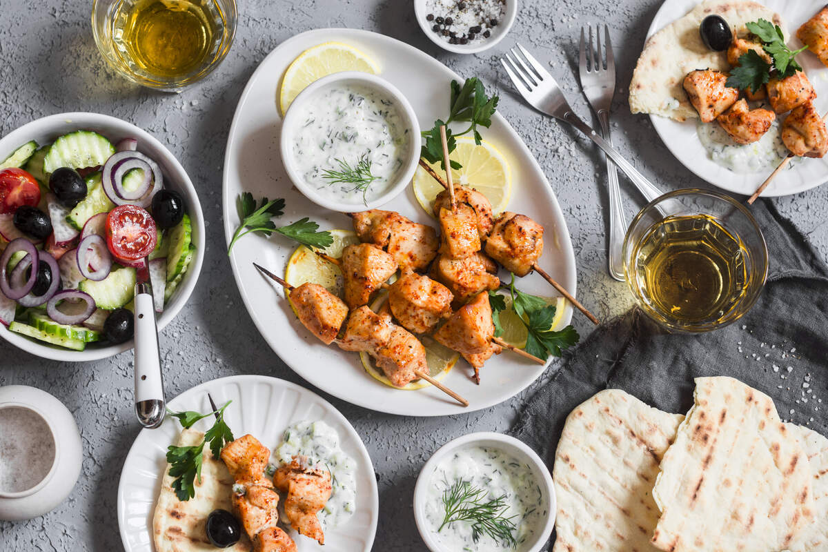 A high-angle overhead shot of a fresh Greek dinner spread arranged on a gray textured background. The centerpiece is a white oval platter filled with seasoned, grilled chicken souvlaki skewers resting on lemon slices and garnished with fresh parsley. Surrounding the platter are several small plates with pita bread and chicken, bowls of creamy tzatziki sauce garnished with dill, two glasses of white wine, and a colorful side salad filled with cucumbers, tomatoes, red onions, and kalamata olives.
