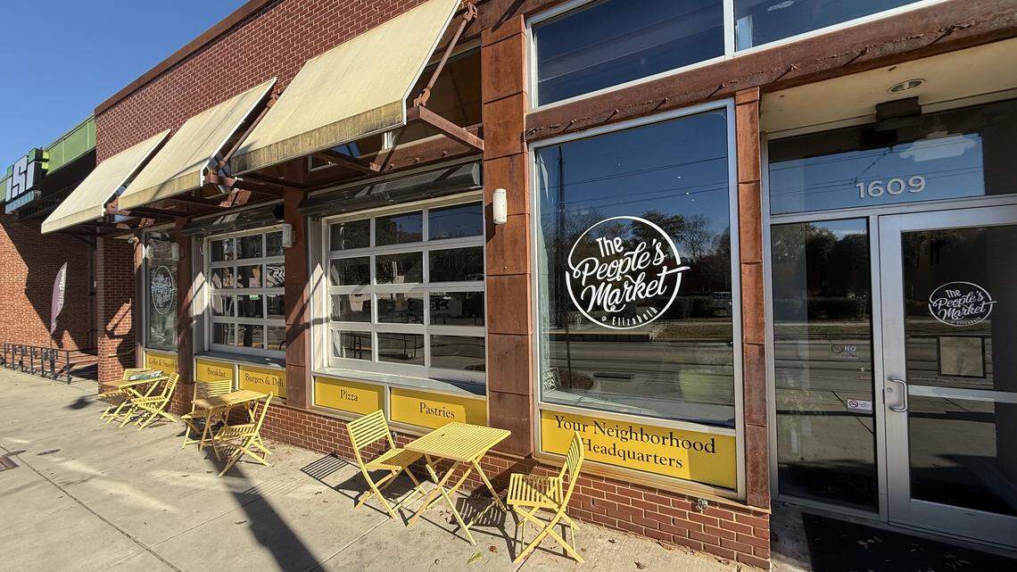 An eye-level, angled shot from the sidewalk of “The People’s Market @ Elizabeth” on a sunny day. The red brick storefront features large windows, glass-paneled garage doors, and weathered tan awnings. Several yellow metal tables and chairs are arranged on the sidewalk for outdoor seating.