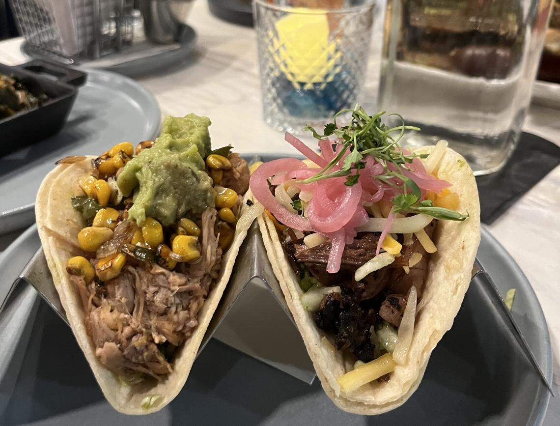 A high-angle, close-up shot of two tacos resting in a metal stand on a gray plate. The taco on the left is filled with shredded pork, roasted corn, and topped with guacamole. The taco on the right contains dark shredded meat, cheese, pickled red onions, and microgreens. Blurred drinking glasses are visible in the background.