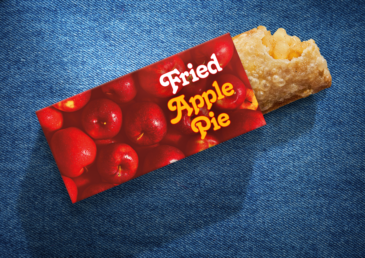A golden-brown, bubbly fried turnovers-style apple pie with a bite taken out of it, revealing the spiced fruit filling inside. The pie is partially tucked into a red cardboard sleeve featuring a pattern of glossy red apples and the words “Fried Apple Pie” written in a stylized white and yellow font. The entire product rests at a diagonal angle against a textured blue denim fabric background.