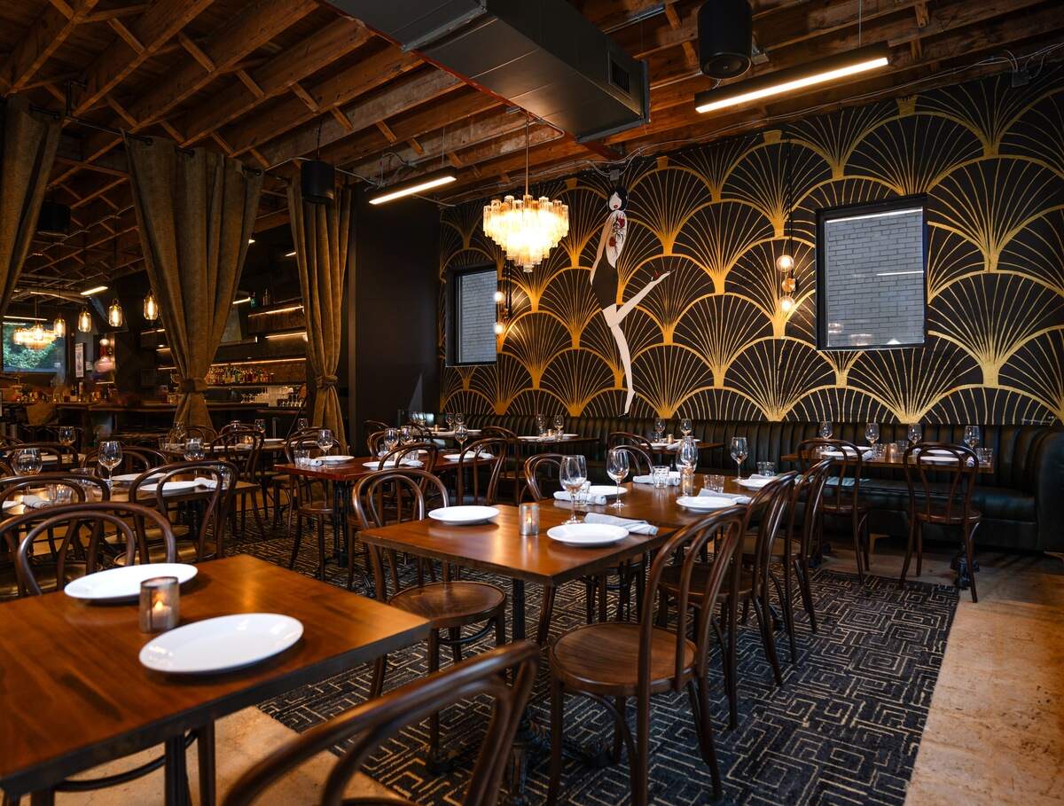 A wide-angle view of a restaurant’s elegant dining room, which blends Art Deco glamour with industrial architecture. A prominent wall features black and gold wallpaper with a large dancer mural. Tables are meticulously set for dinner under a high, exposed-rafter ceiling from which a crystal chandelier hangs.
