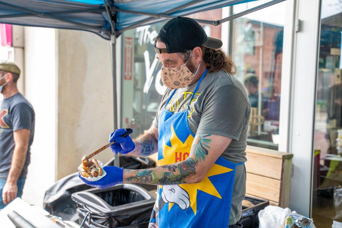 A vendor from “Vic The Chili Man” is preparing a hot dog under a blue tent. The man, with long hair and a beard, is wearing a black baseball cap backward, a gray t-shirt, and a bright blue apron with a yellow star and hand graphic. He has a patterned face mask on and is wearing blue gloves as he uses a spoon to add toppings to a hot dog in a paper tray. Tattoos are visible on his arms. A customer in a gray t-shirt and baseball cap stands in the background.
