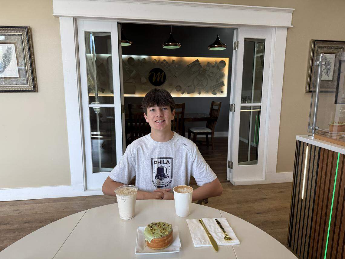 A teenager sits at a white circular table in the cafe. In front of him are two coffee drinks and a pistachio-topped pastry. Behind him, white sliding doors are open, revealing the private dining room with the backlit kitchen utensil wall art.