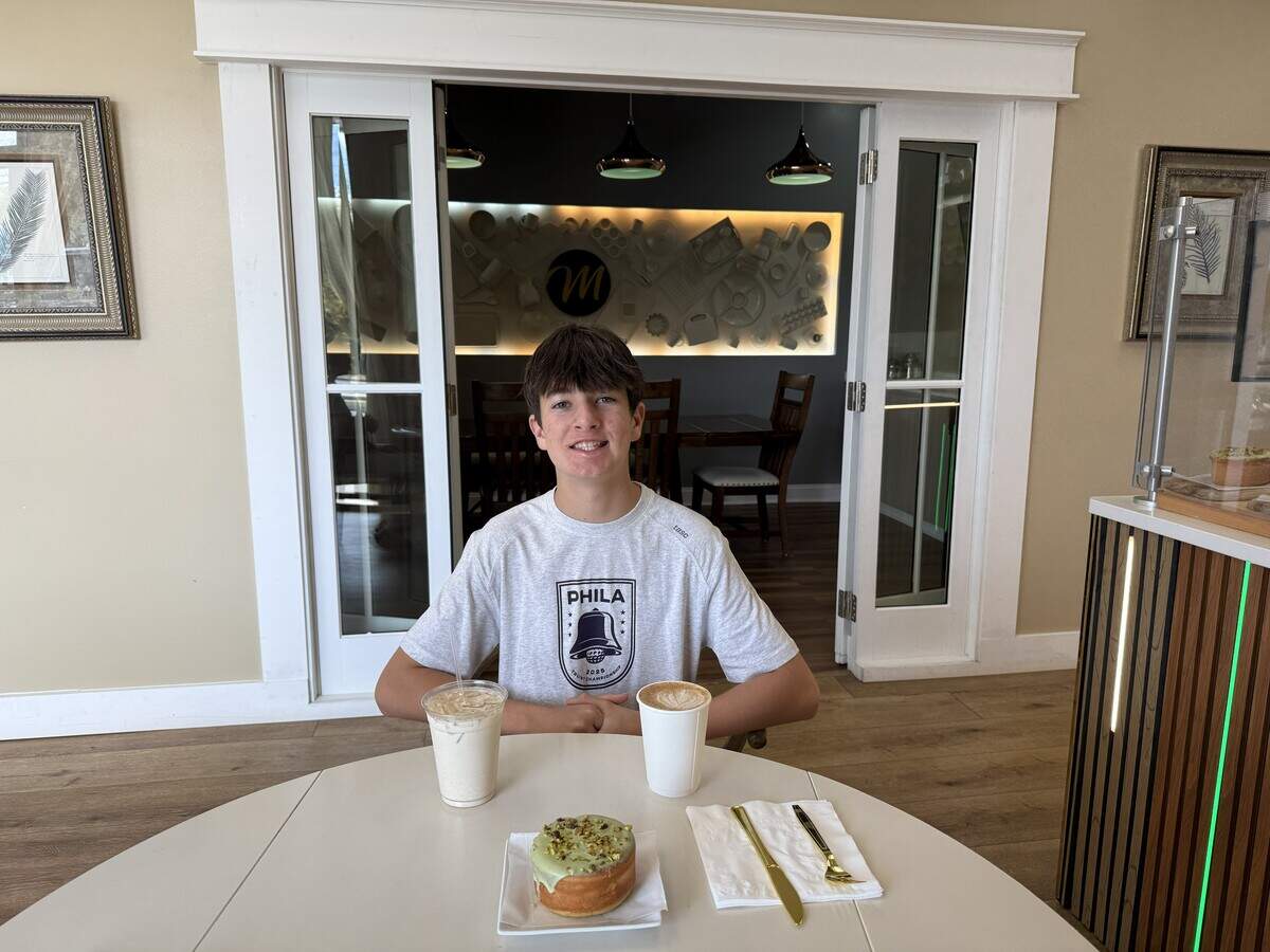 A teenager sits at a white circular table in the cafe. In front of him are two coffee drinks and a pistachio-topped pastry. Behind him, white sliding doors are open, revealing the private dining room with the backlit kitchen utensil wall art.