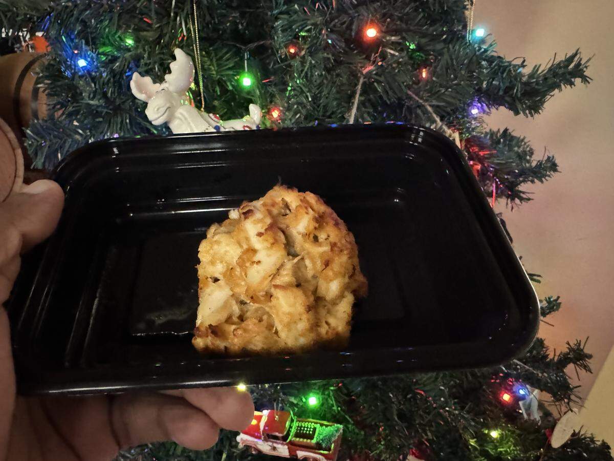 A single crab cake served in a black takeout container held in front of a decorated Christmas tree.