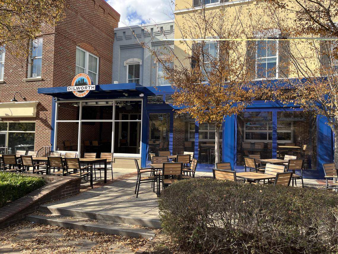 An eye-level, wide shot shows the outdoor patio of the Dilworth restaurant on a sunny day. The patio is filled with numerous empty wooden tables and chairs set on a stone terrace. A large blue awning structure extends from the restaurant, with a circular “DILWORTH” logo visible on the corner, set against the red brick and yellow-painted buildings. Trees with yellowing autumn leaves are in the foreground.
