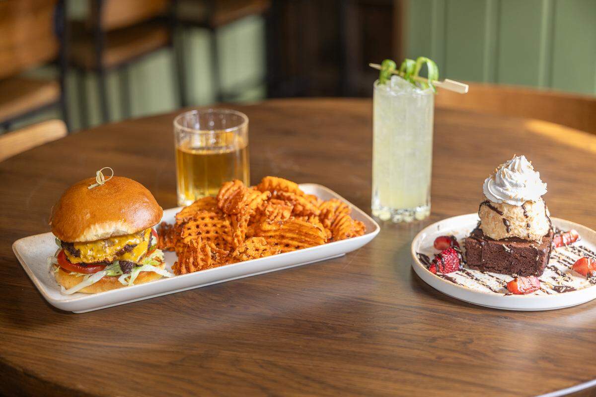 A wooden tabletop features a spread of pub favorites. In the foreground, a double cheeseburger with lettuce and tomato is served on a rectangular white plate alongside a generous pile of seasoned waffle fries. Behind the plate sits a short glass of amber-colored beer and a tall, refreshing pale yellow cocktail garnished with a sophisticated cucumber ribbon. To the right, a decadent brownie dessert completes the spread.