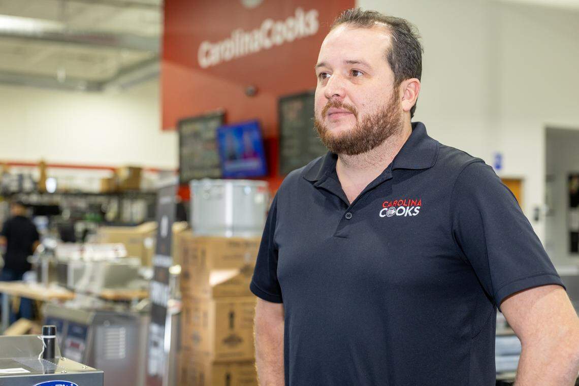A medium, eye-level shot of a business owner with a beard, wearing a black polo shirt with the “Carolina Cooks” logo, standing inside a commercial kitchen supply warehouse. The business owner is looking off to the side, and the background is blurred with stacks of boxes and stainless steel equipment.
