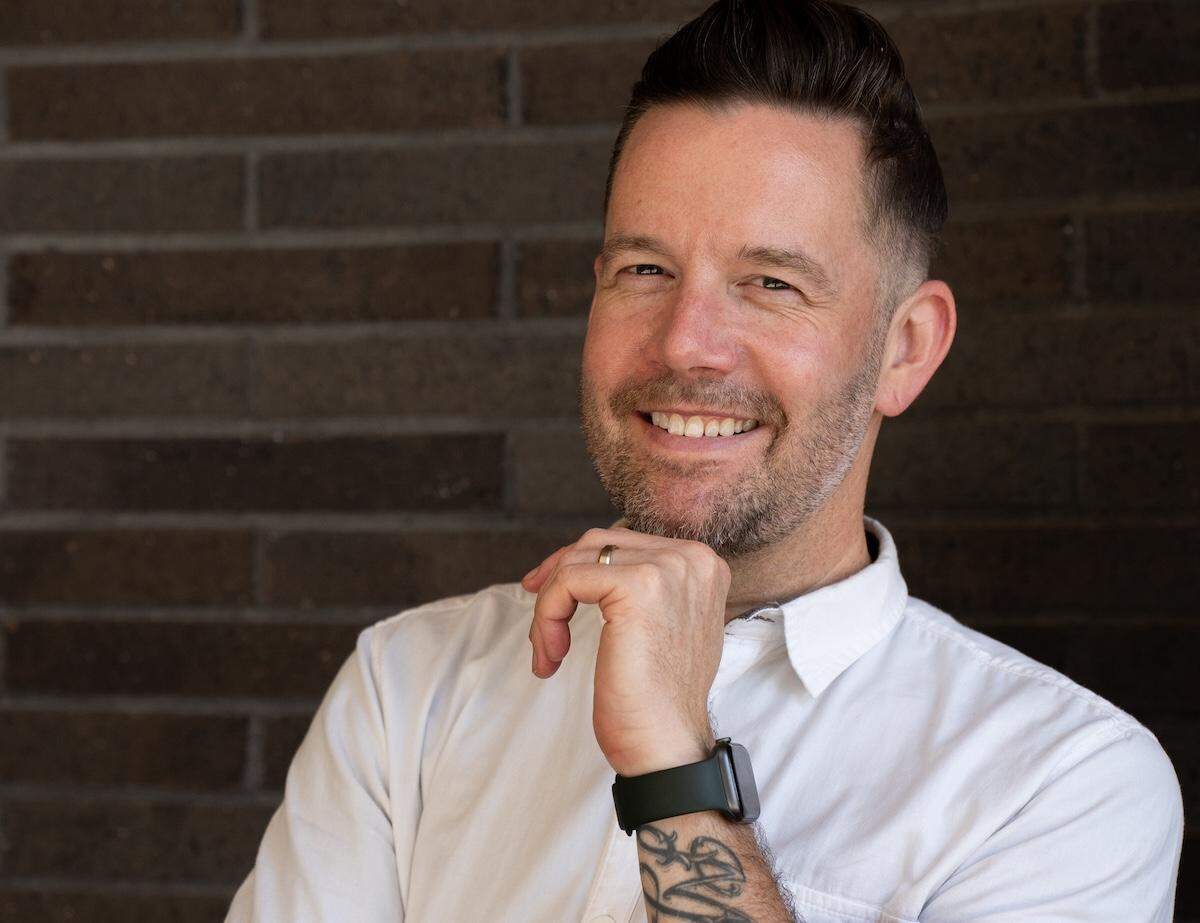 Headshot of a smiling chef, wearing a white shirt and standing against a dark brick wall.