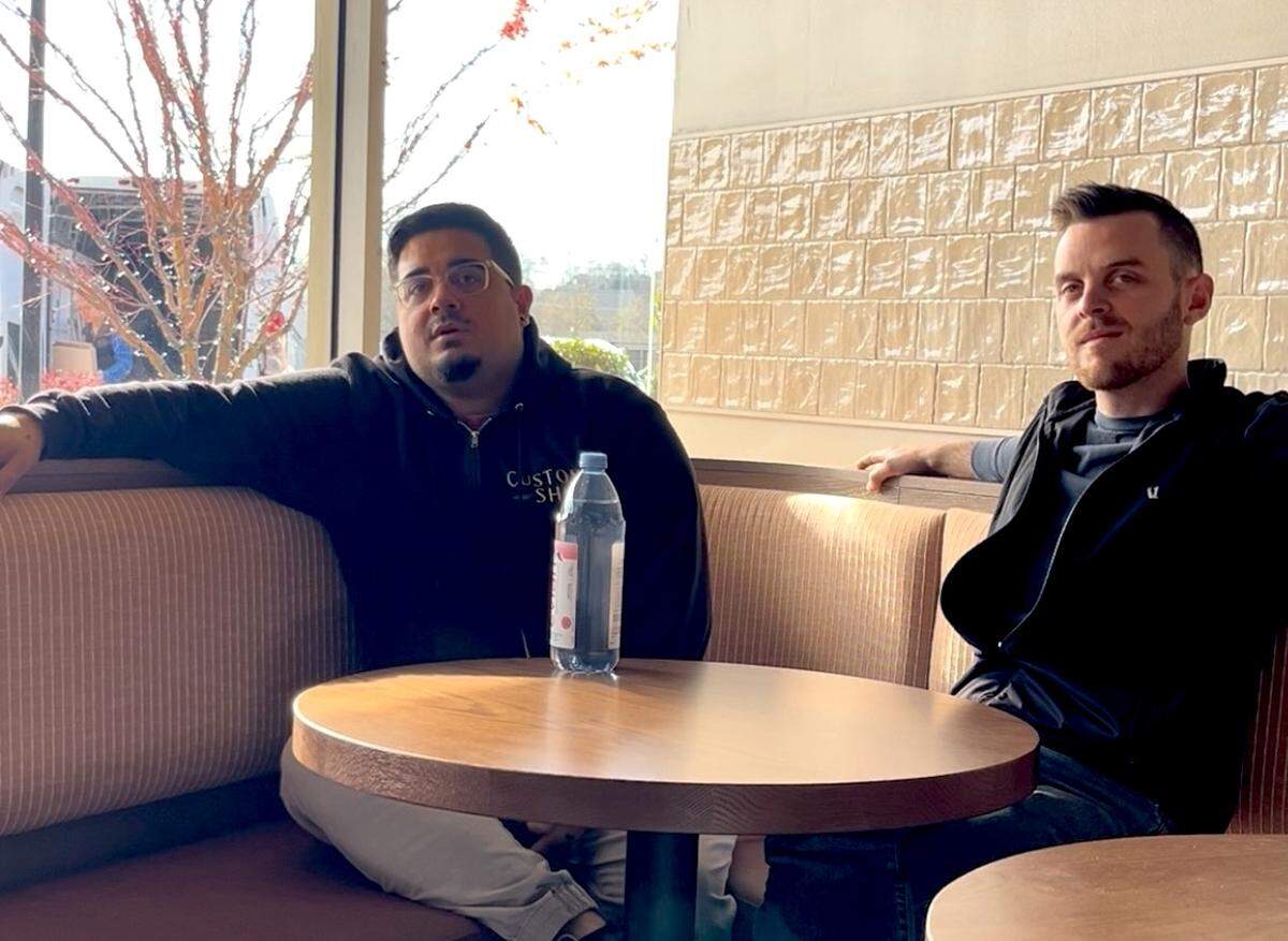 Two restaurateurs are seated across a small, round wooden table in a booth inside a brightly lit restaurant or cafe. The one on the left wears a dark hoodie, and the one on the right wears a black jacket and vest. The background shows a large window with bare tree branches and bright sunlight, and the interior wall behind the man on the right has textured, light-colored square tiling.”