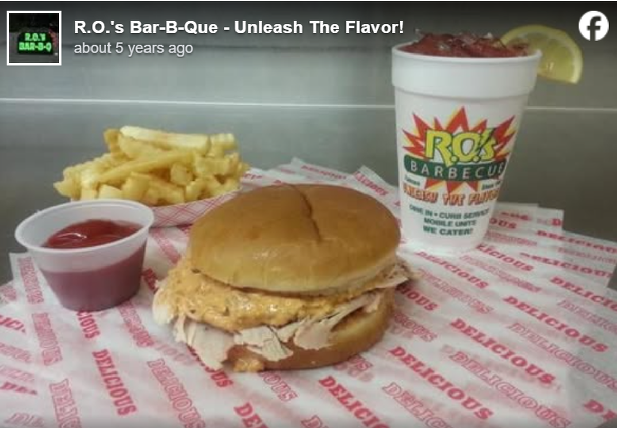 A barbecue sandwich filled with sliced meat and a creamy orange sauce sits on a branded paper liner that says “DELICIOUS.” Behind the sandwich is a side of crinkle-cut fries, a small cup of ketchup, and a large styrofoam cup featuring the “R.O.’s Barbecue” logo and a lemon wedge on the rim.