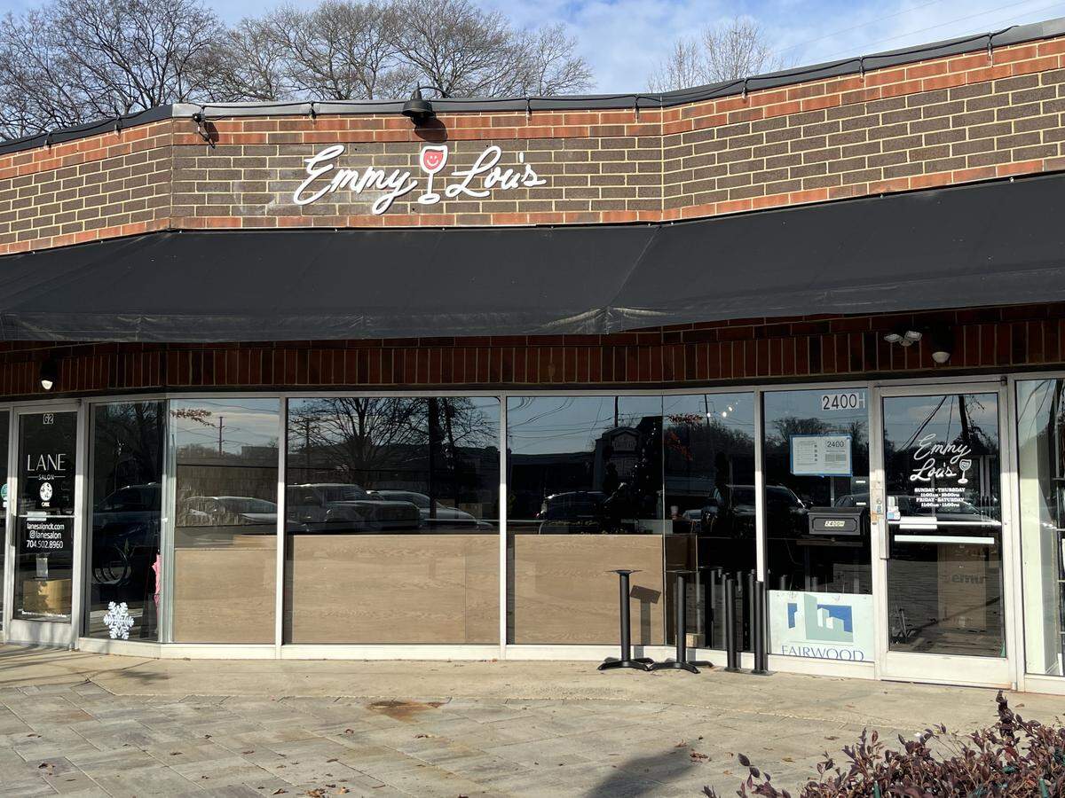 A photograph of the storefront for ‘Emmy Lou’s,’ a business located in a brick strip mall. The name is written in white cursive with a wine glass graphic above a black awning. Large front windows reflect the parking lot and sky.