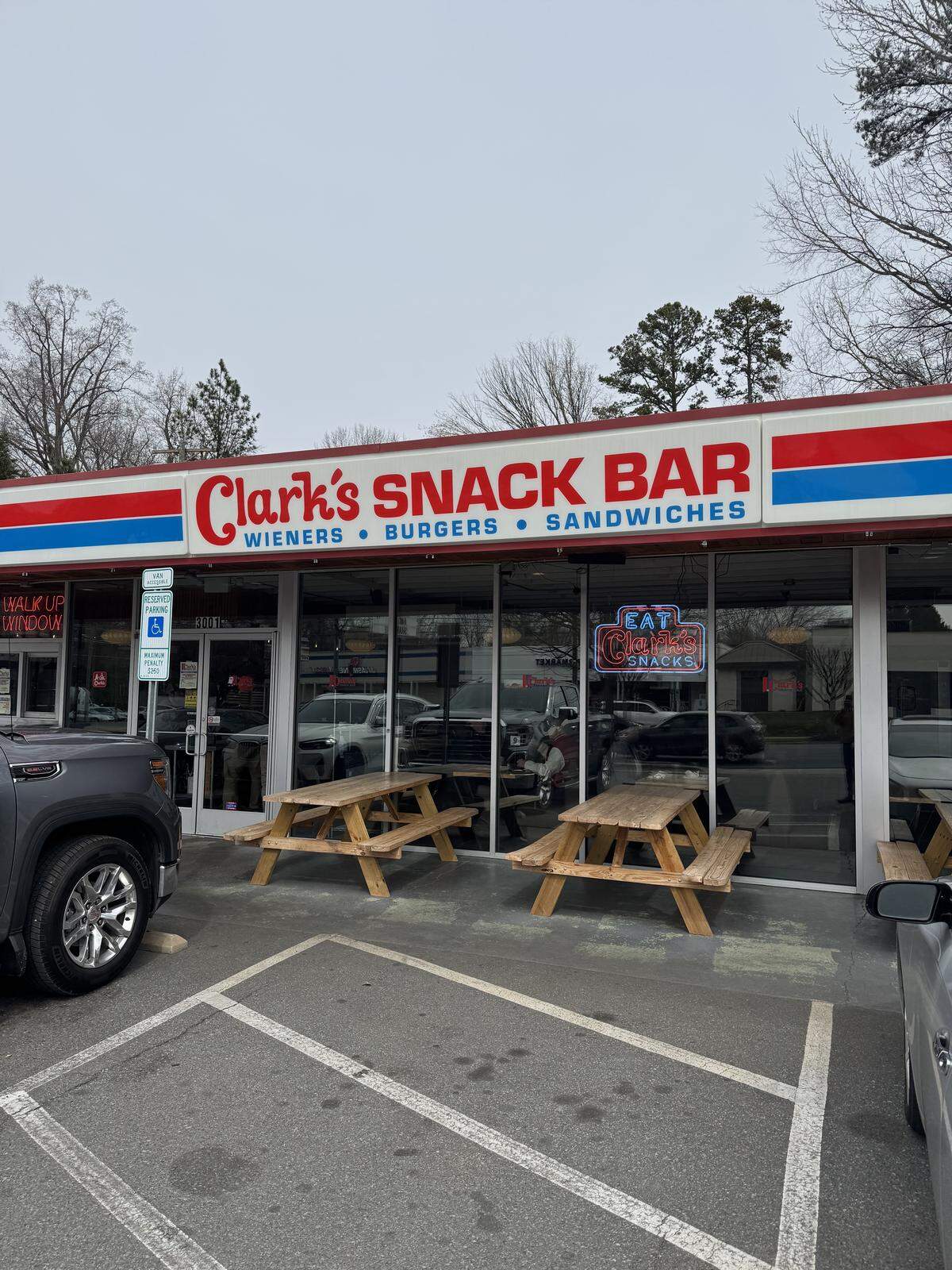 A wide exterior shot of Clark’s Snack Bar, a one-story restaurant with a red, white, and blue striped awning. Two wooden picnic tables sit on the sidewalk in front of large glass windows.