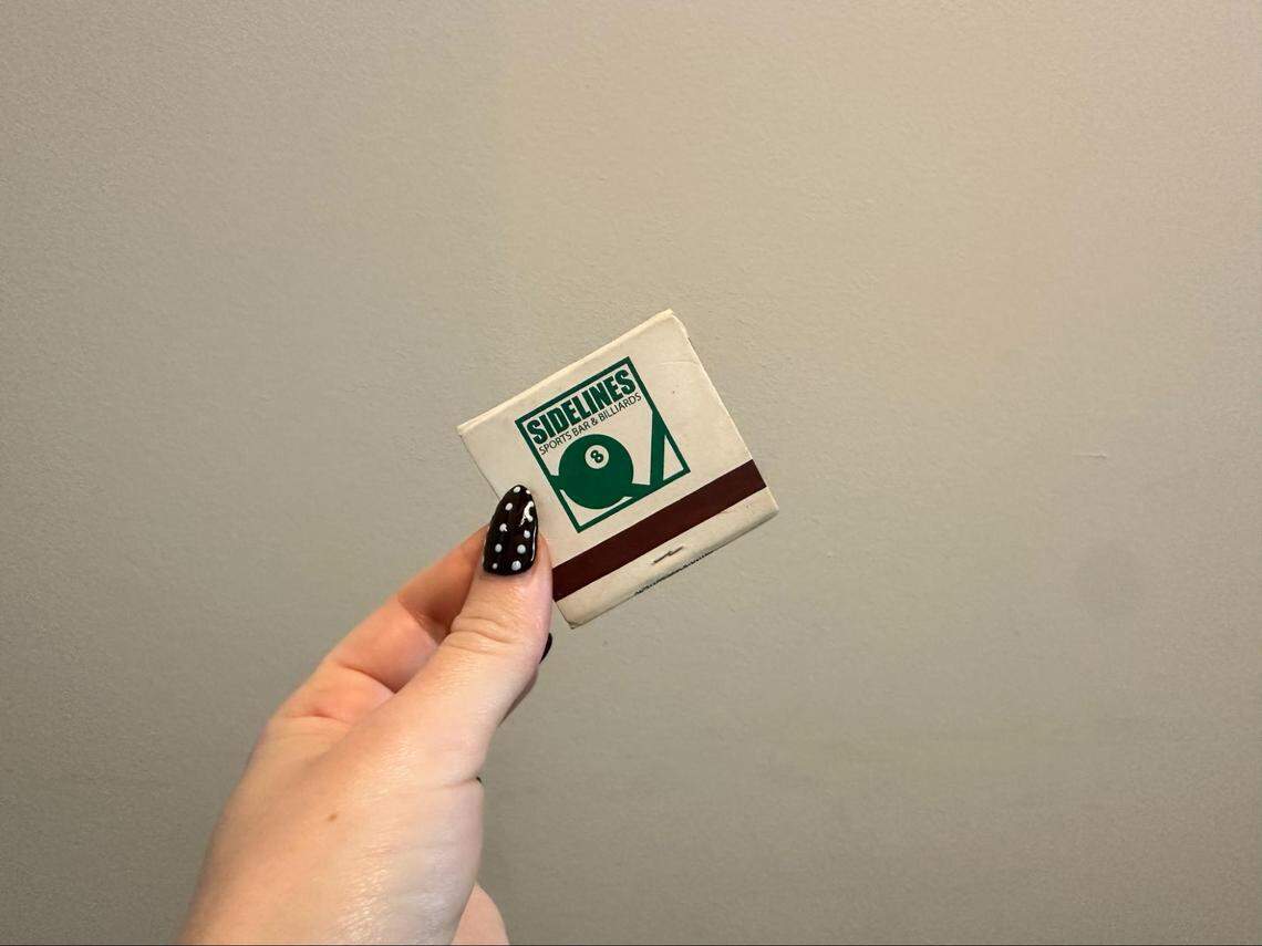 A person’s hand, with a black and white polka-dot manicure, holds up a matchbook against a plain gray wall. The matchbook is for “Sidelines Sports Bar & Billiards” and features its green 8-ball logo.