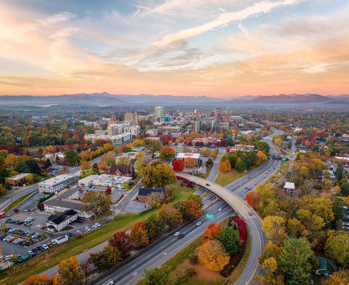 A wide, elevated shot of Asheville, NC, nestled in the mountains during fall. The foreground features a curving highway overpass amidst trees with brilliant red and orange leaves, leading towards the downtown area under a pastel-colored sky.
