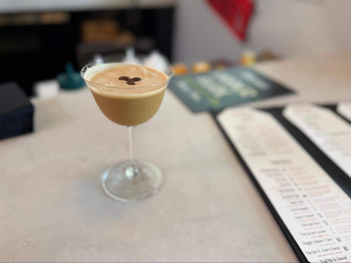 A close-up, eye-level shot of an espresso martini in a coupe glass, resting on a light-colored tabletop. The drink has a creamy, tan-colored foam top and is garnished with three coffee beans arranged in the center. The cocktail is in sharp focus, while the background is blurred, showing menus laid out on the table.