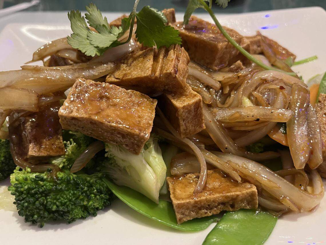 A close-up of a stir-fried dish plated on a white square plate. The dish features golden-brown fried cubes of tofu tossed with caramelized onions and a light brown, glossy sauce. It is served over vibrant green vegetables, including broccoli florets and snow peas, and garnished with fresh cilantro leaves.