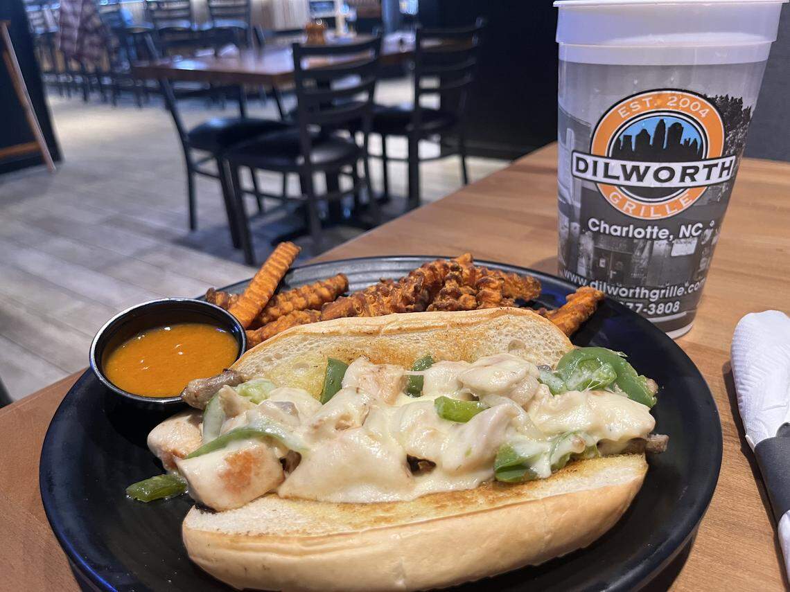 A high-angle, close-up shot of a meal on a wooden table. A black plate holds a cheesesteak sandwich, topped with meat, green peppers, and melted white cheese, alongside a serving of sweet potato waffle fries and a small ramekin of orange dipping sauce. To the right is a large “Dilworth Grille” drink cup.