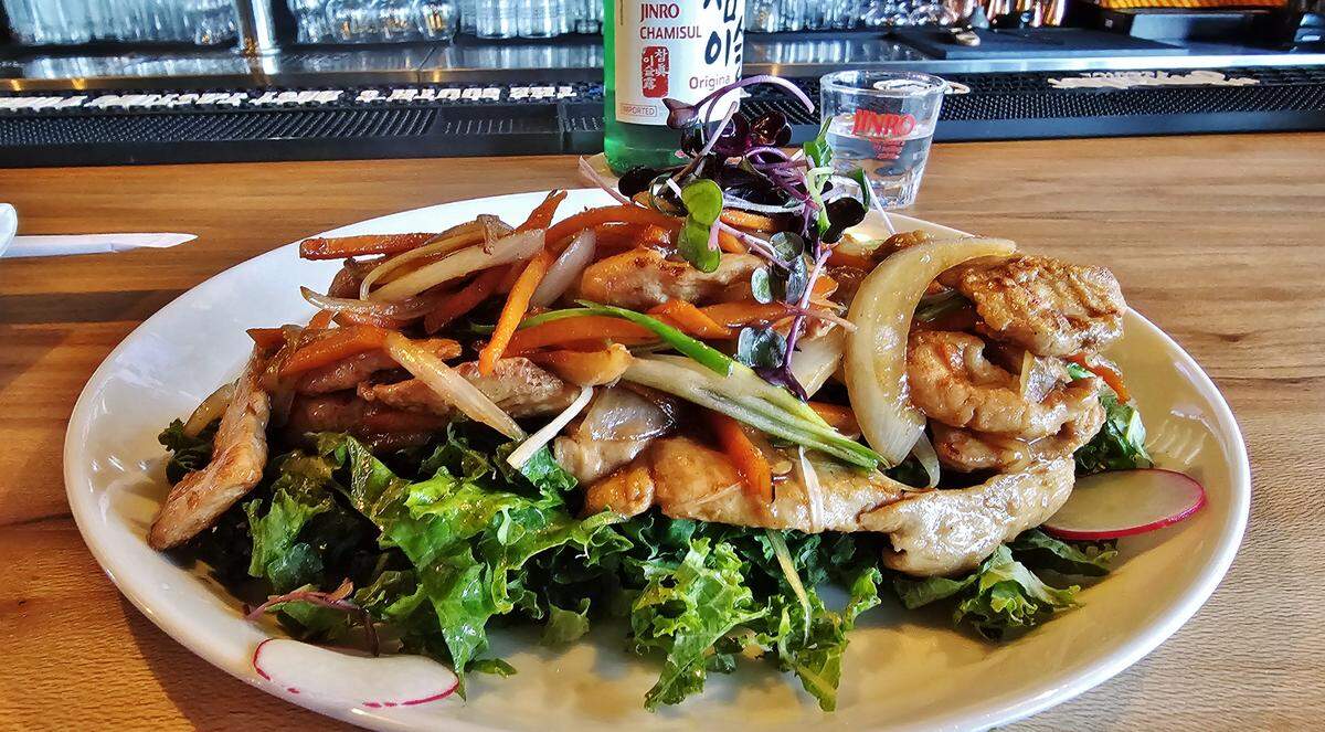 A beautifully plated Korean dish on a white plate, sitting on a wooden table. The dish features stir-fried meat mixed with sliced onions, carrots, and scallions. It is served on a bed of fresh, leafy greens and garnished with microgreens. In the background, a bottle of soju and a shot glass are visible.