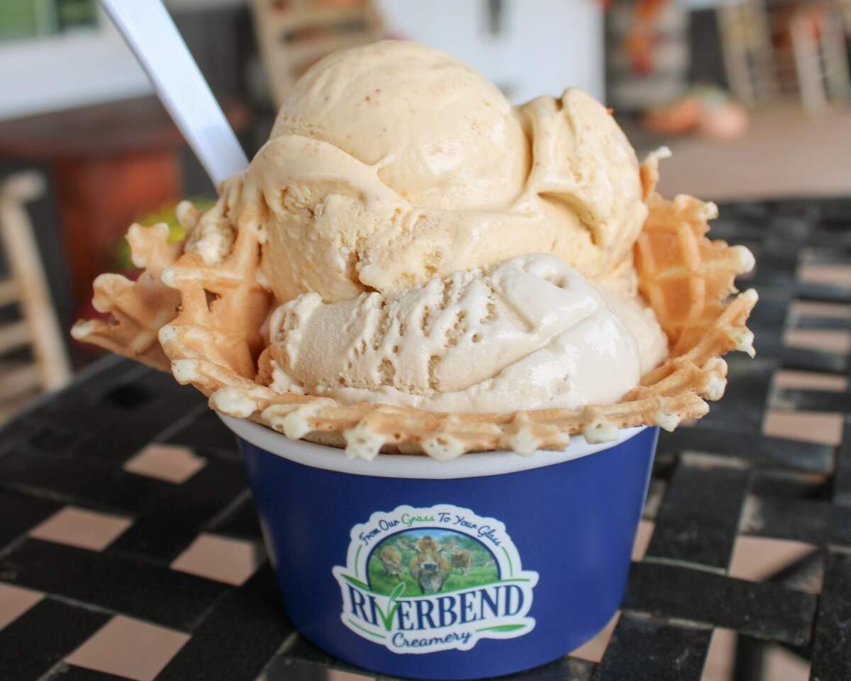 A close-up shot of two large scoops of creamy, tan-colored ice cream served in a waffle cone bowl. The waffle bowl sits inside a blue paper cup with the “Riverbend Creamery” logo, which features an image of two cows in a pasture. The cup is resting on a black mesh patio table, and a white spoon is sticking out from behind the ice cream.