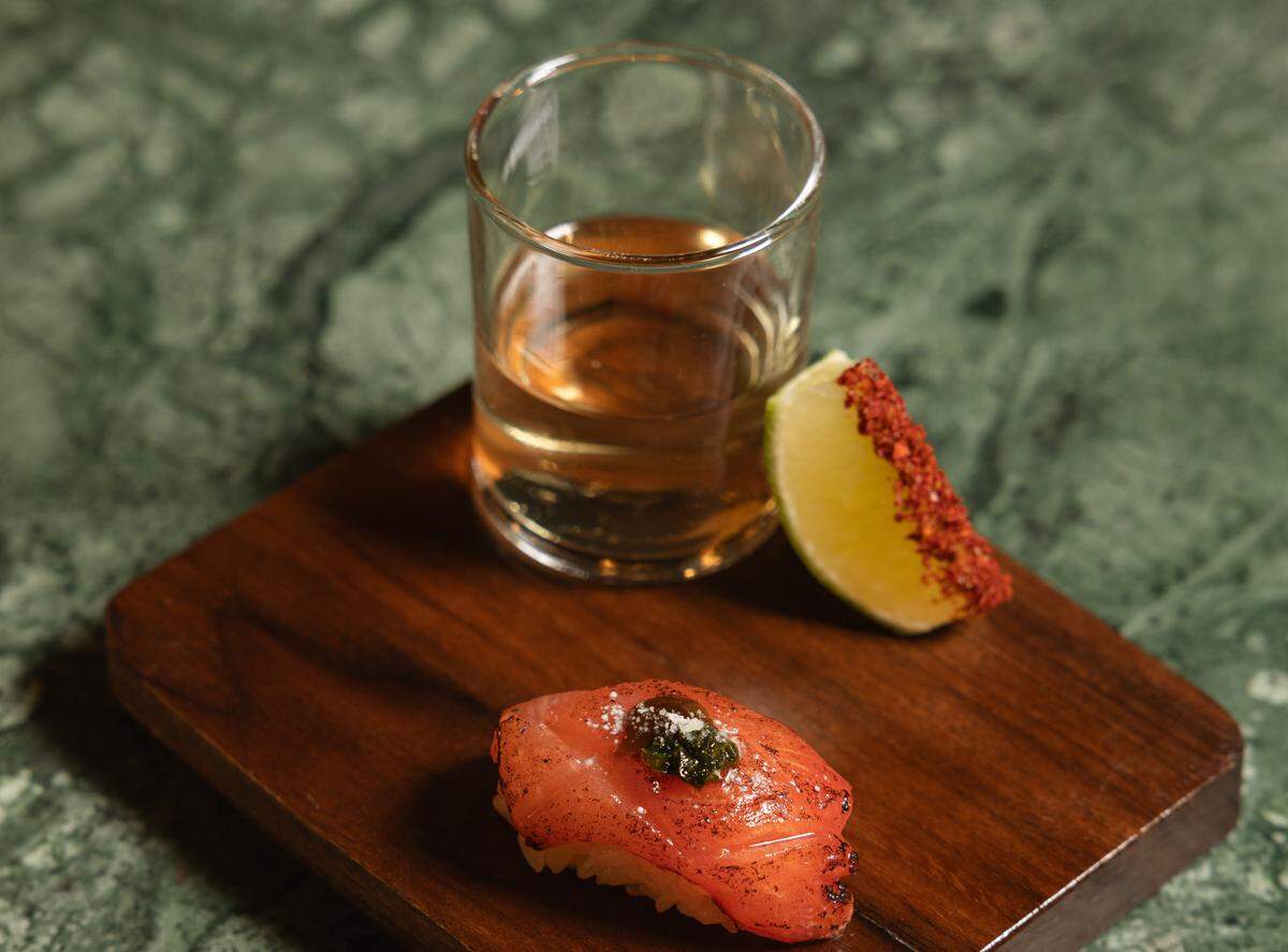 A close-up shot of a small wooden serving board on a green marble surface. On the board is a single piece of torched nigiri sushi topped with a dark garnish, a small glass filled with an amber-colored liquid, and a lime wedge coated on one side with red chili seasoning.