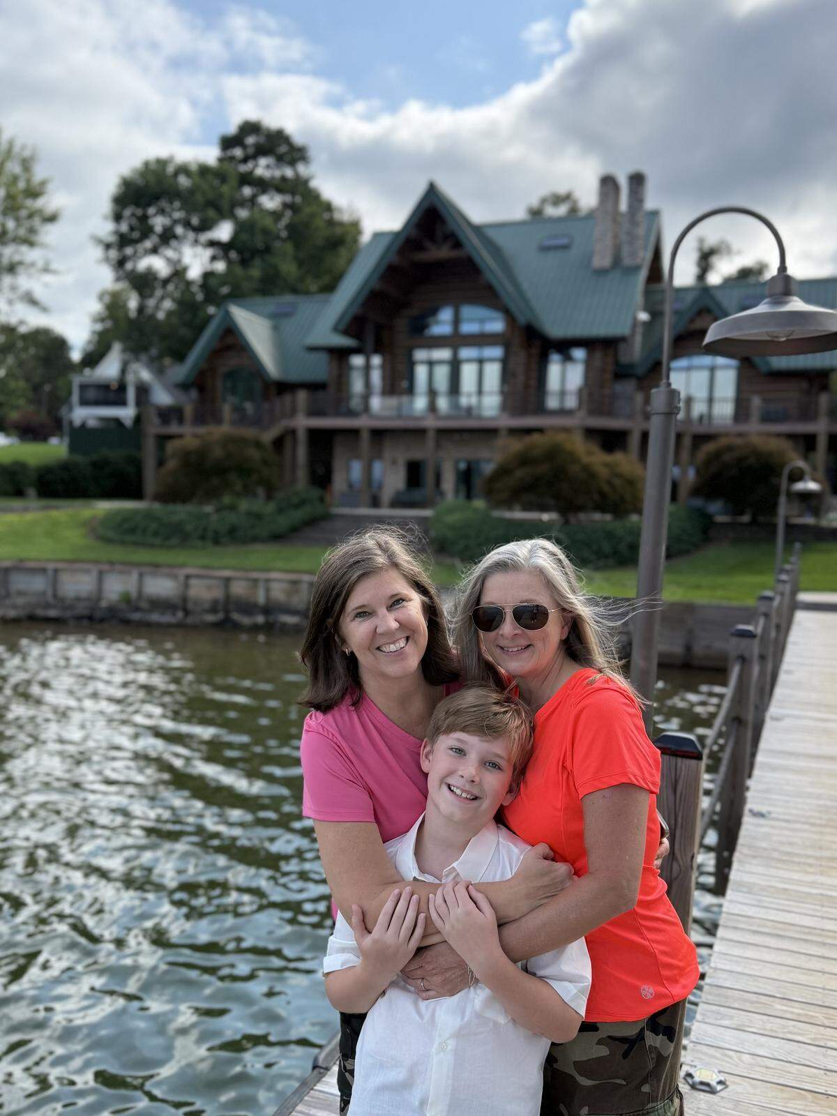 Three family members, two adults and a child, smile and hug for a photo on a wooden dock. The large log cabin is visible in the background across the water.