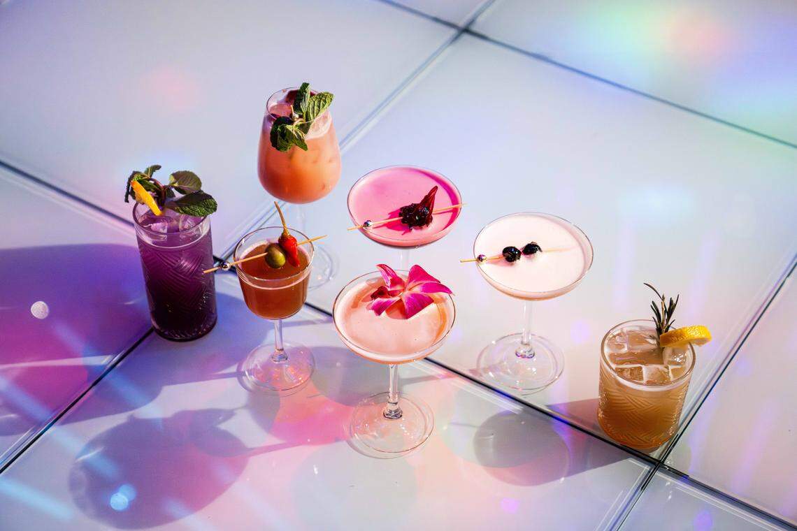 A colorful collection of seven unique cocktails arranged on a light-colored, tiled floor. The scene is illuminated by vibrant, multi-colored club lighting in shades of purple, blue, and pink. The drinks vary in glass style and garnish, featuring elements like fresh mint, edible orchids, dark cherries, and rosemary sprigs, creating a festive and high-energy atmosphere.