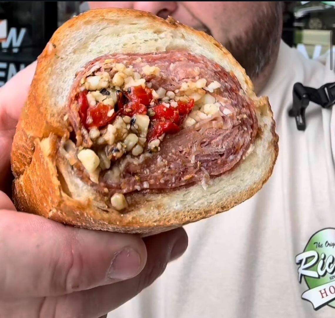 A close-up shot of a person holding a large Italian sub sandwich that has been sliced to show the end. The sandwich is densely packed with many layers of thinly sliced deli meat rolled tightly. In the center of the meat roll is a generous filling of chopped garlic, red peppers, and herbs. The crusty bread roll is golden brown and slightly dusted with flour.
