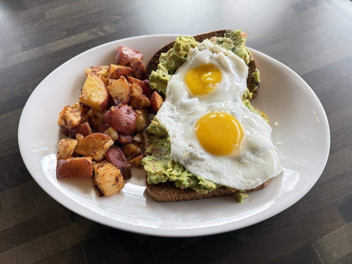 A close-up of a white plate featuring a slice of toast topped with mashed avocado and two sunny-side-up eggs, served alongside a pile of seasoned, roasted red potatoes.