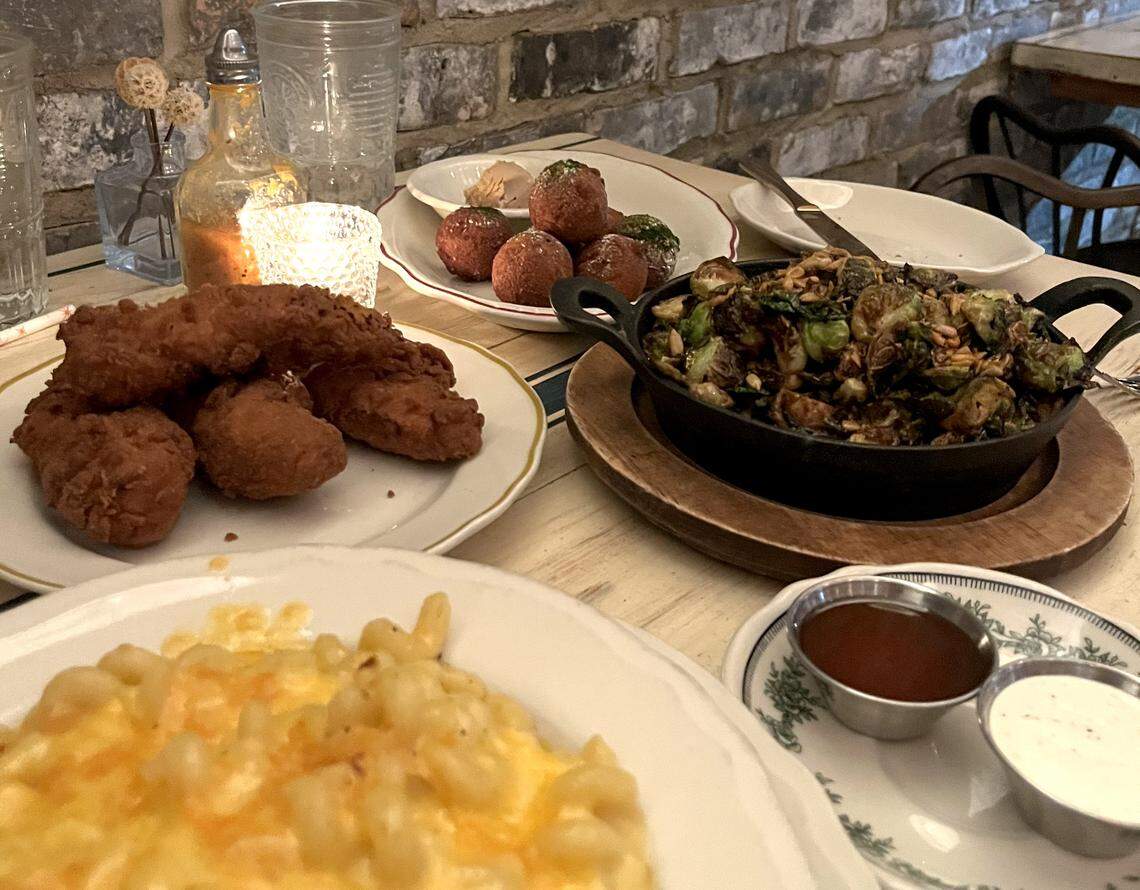 An eye-level, medium shot of a dinner spread on a light wooden table against a rustic brick wall. The meal includes a large plate of crispy, deep-fried chicken tenders in the foreground, a bowl of creamy, baked macaroni and cheese, a small cast-iron skillet filled with charred Brussels sprouts topped with toasted seeds, a splate featuring several fried dough balls or croquettes served with a small dollop of butter and two small metal ramekins containing dipping sauces (one dark red, one white). The table is lit by a small, flickering white candle in a textured glass holder, creating a warm and cozy dining atmosphere.