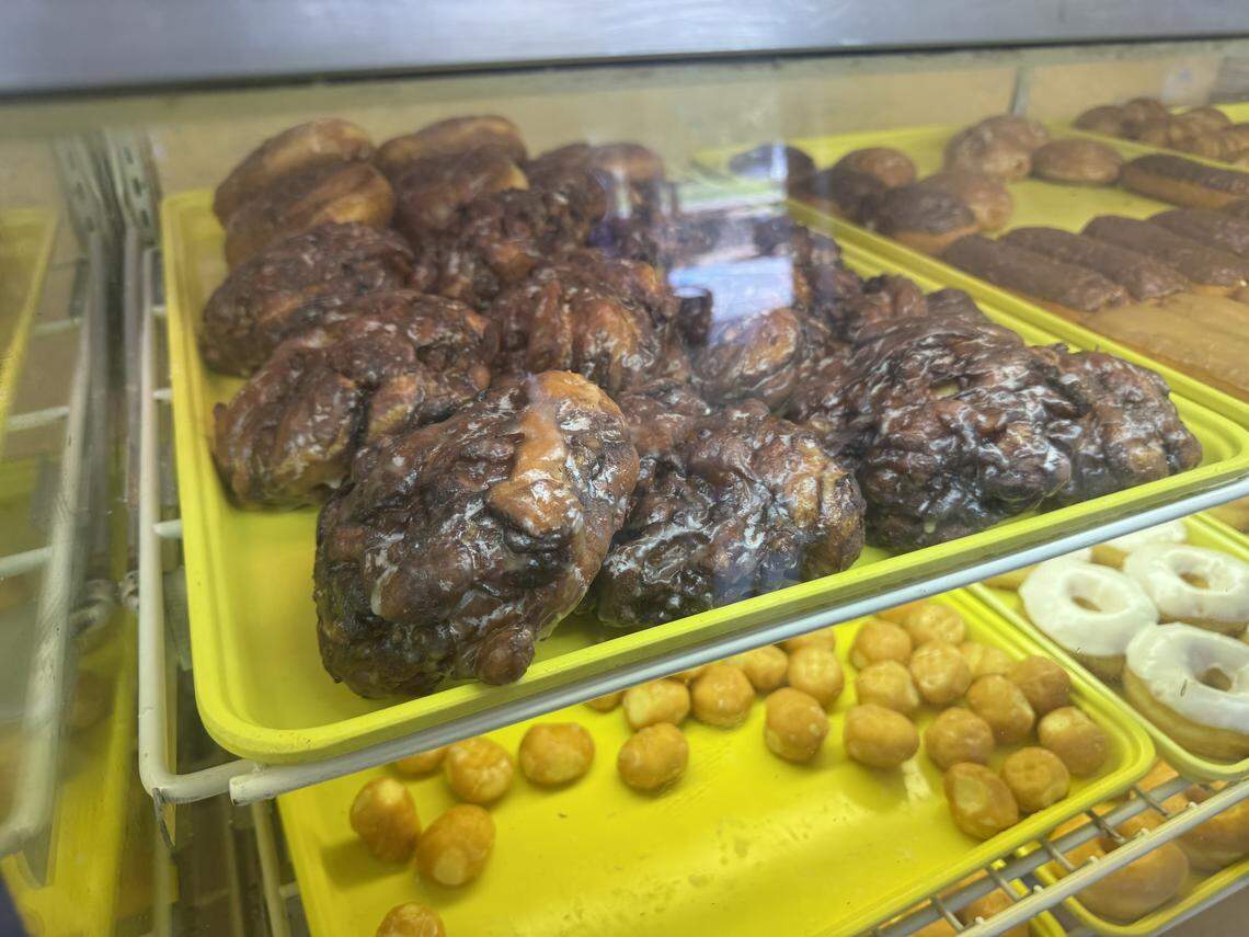 A close-up, angled shot into a glass display case at a donut shop. The focus is on a yellow tray laden with large, lumpy apple fritters covered in a clear glaze. A lower shelf holds a tray with golden donut holes and white-iced donuts.