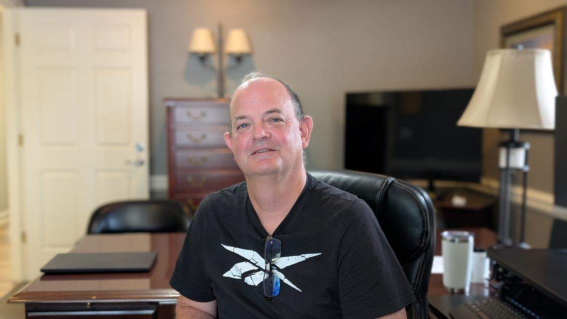 A person with short hair, wearing a black V-neck t-shirt with a white graphic, sitting in a black leather office chair at a large wooden desk. They are looking at the camera, and glasses are hanging from their collar. The background shows a professional office setting with a TV, lamp, and wooden cabinet.