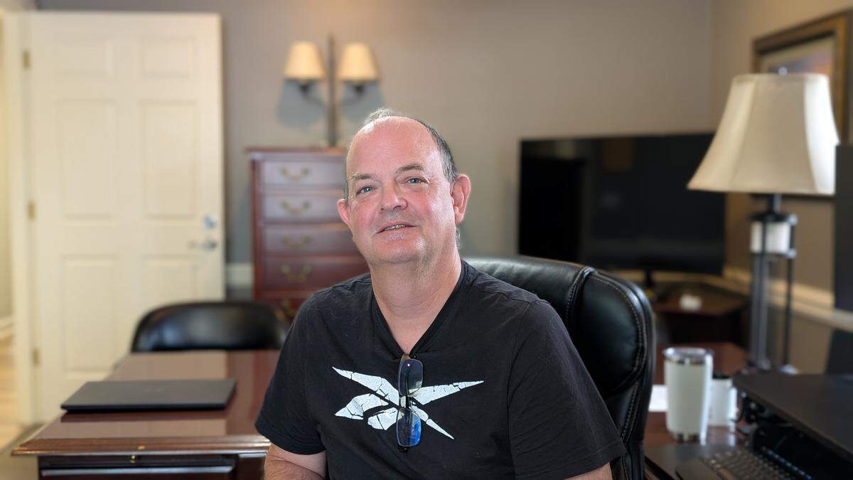 A person with short hair, wearing a black V-neck t-shirt with a white graphic, sitting in a black leather office chair at a large wooden desk. They are looking at the camera, and glasses are hanging from their collar. The background shows a professional office setting with a TV, lamp, and wooden cabinet.