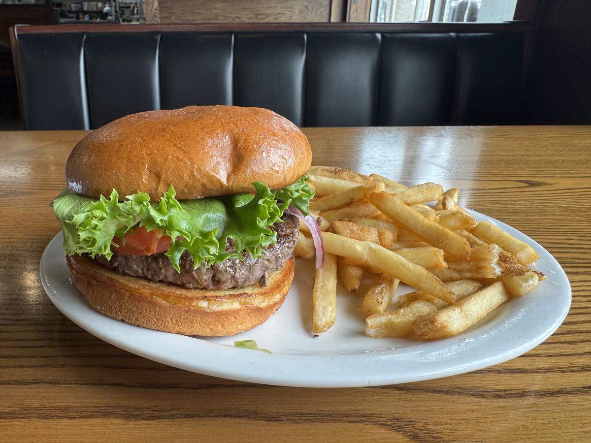 A close-up of a juicy cheeseburger with fresh green lettuce, tomato, and red onion on a toasted brioche bun, served with a side of golden-brown french fries on a white plate.