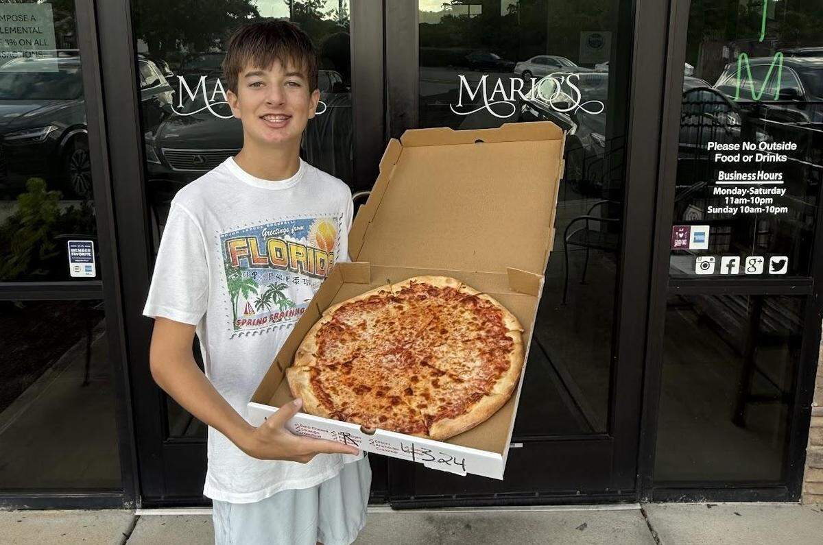 A young teenage boy with brown hair and braces, wearing a white t-shirt with a “Greetings from Florida” graphic, stands outside the entrance of a restaurant named “Mario’s.” He is smiling proudly while holding a large, open pizza box containing a whole, round cheese pizza. The restaurant’s glass door and a sign with its logo and business hours are visible behind him. The pizza is a classic cheese pizza with a bubbly, golden-brown top.
