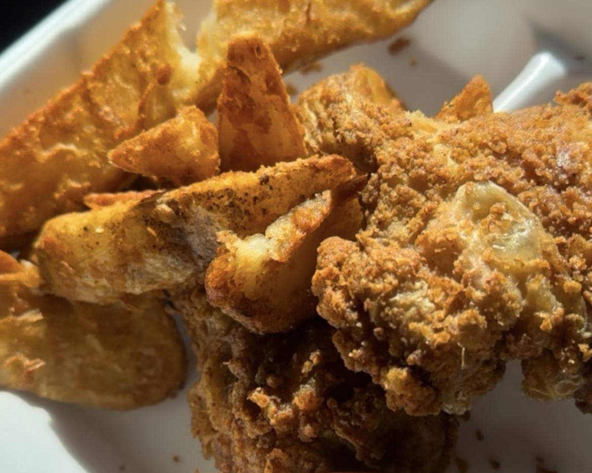 A tight, extreme close-up shot focusing on the textures of golden-brown, crispy fried chicken pieces and seasoned potato wedges, all packed together in a white to-go container under bright, direct light.
