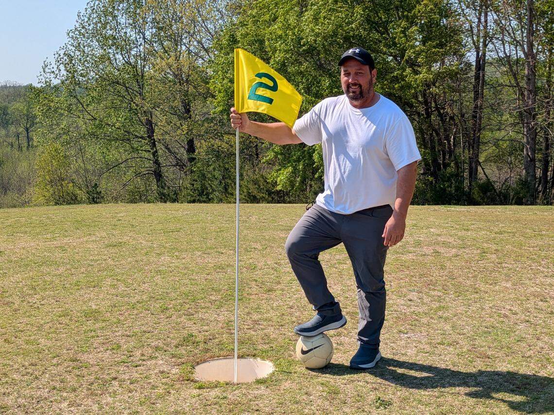 A person with a beard and cap stands triumphantly on a golf green next to an oversized hole, holding the pin of a yellow flag marked with a “2” and rests a sneaker on a full-sized soccer ball. The background shows green trees against a clear sky.