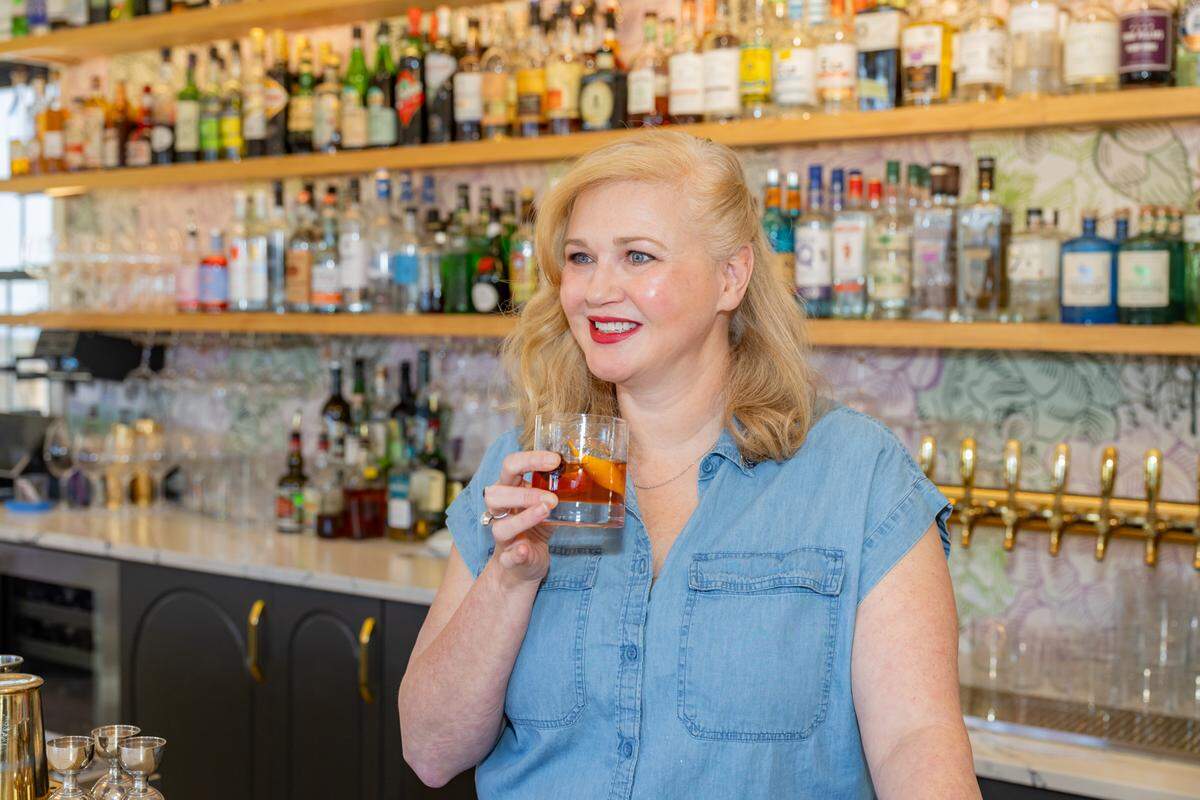 A smiling blonde woman in a denim shirt holds a short glass with a drink, possibly a cocktail with an orange peel, in a well-stocked bar. She is looking to her right, with a pleasant expression and red lipstick. Behind her, multiple shelves are filled with various bottles of liquor, and a marble countertop with bar equipment and taps is visible. The background is slightly blurred, emphasizing the woman.